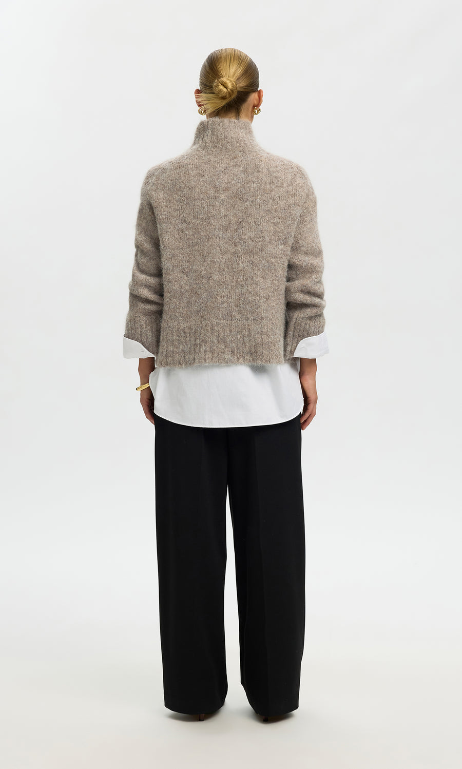 Plümo  Individual with a bun, in the Beel Rollneck beige sweater, white shirt, and black pants, stands facing away.