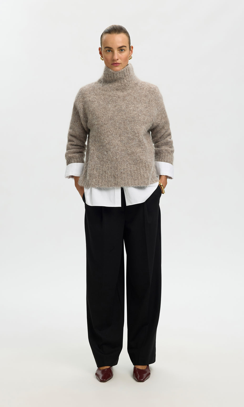 Plümo  Person in the Beel Rollneck beige knit, white shirt, black wide-leg pants, and brown shoes stands facing forward.