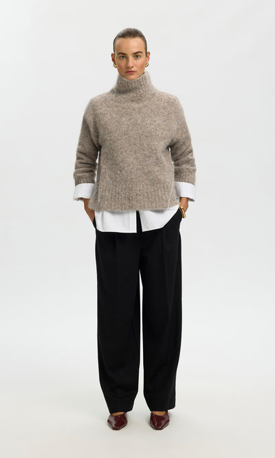 Plümo  Person in the Beel Rollneck beige knit, white shirt, black wide-leg pants, and brown shoes stands facing forward.