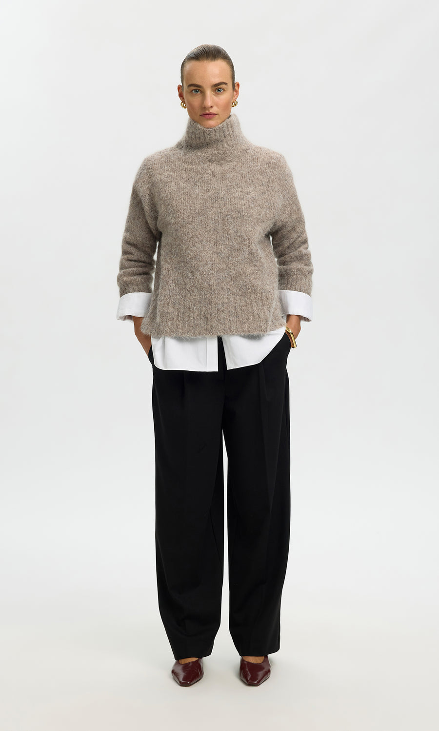 Plümo  Person in the Beel Rollneck beige knit, white shirt, black wide-leg pants, and brown shoes stands facing forward.