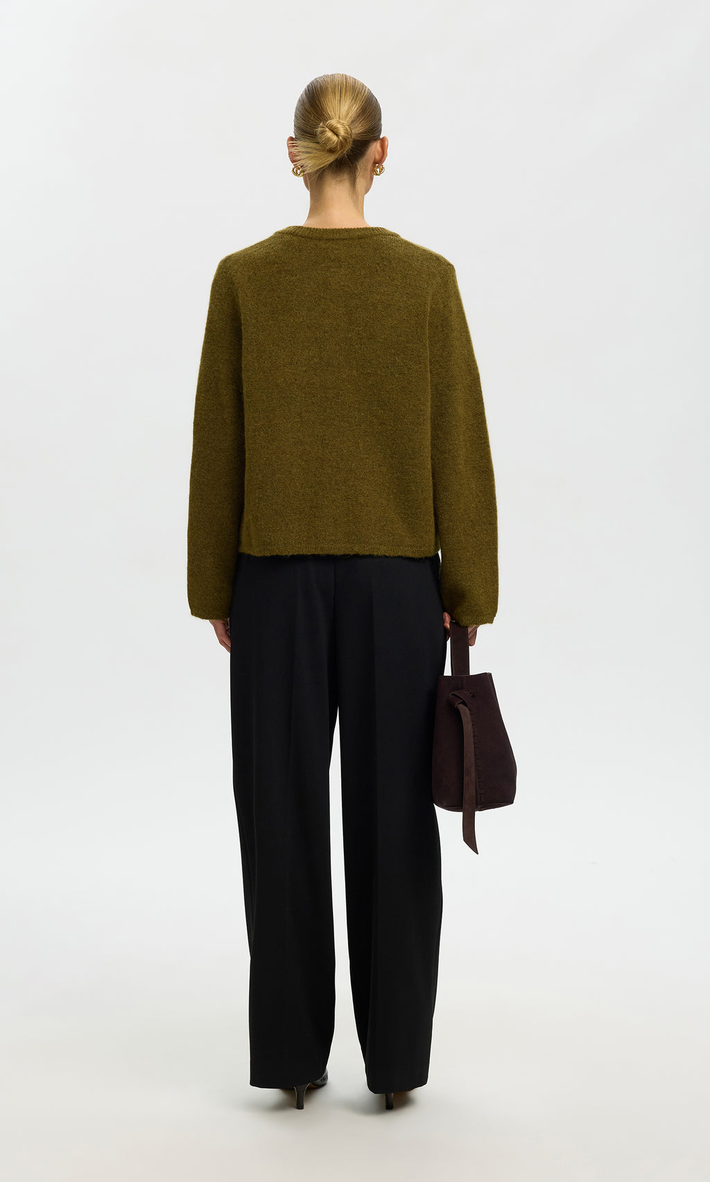 Plümo  Person with blonde hair in a bun, seen from behind in the Lucinda Cardigan moss green and black pants, holding a brown bag.