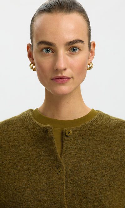 Plümo  Woman with slicked-back hair and gold hoops wears the Lucinda Cardigan in moss green, looking directly at the camera.