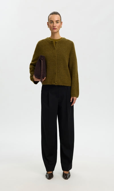 Plümo  Person in a moss green Lucinda Cardigan, black pants, and brown shoes holds a folded dark item against a plain background.