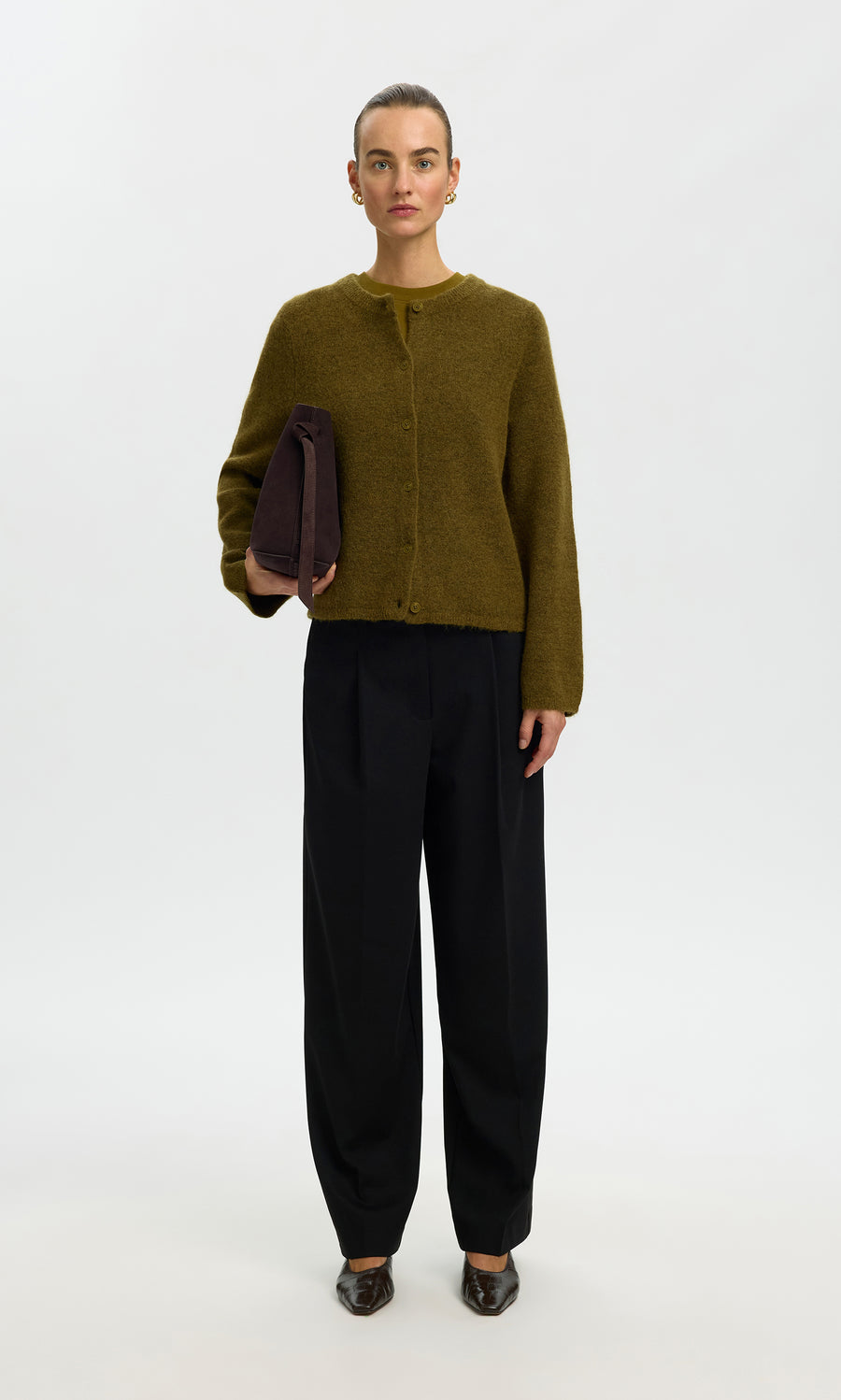 Plümo  Person in a moss green Lucinda Cardigan, black pants, and brown shoes holds a folded dark item against a plain background.