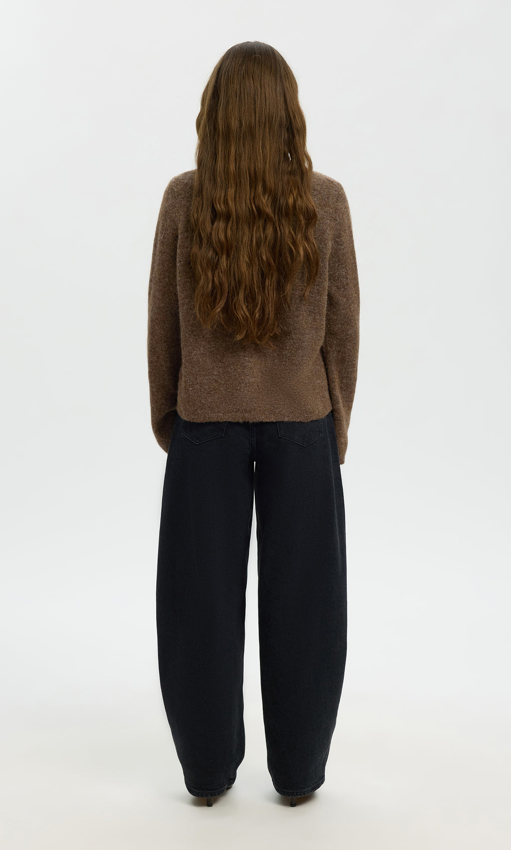 Plümo  Person with long wavy brown hair wearing the Findlay Cardigan and loose black pants, standing facing away.