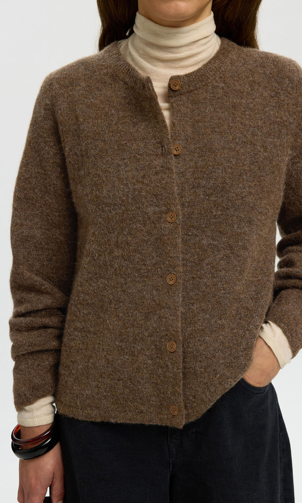 Plümo  Person in the Findlay Cardigan, an airy brown knit crew neck, over a cream turtleneck and black pants.