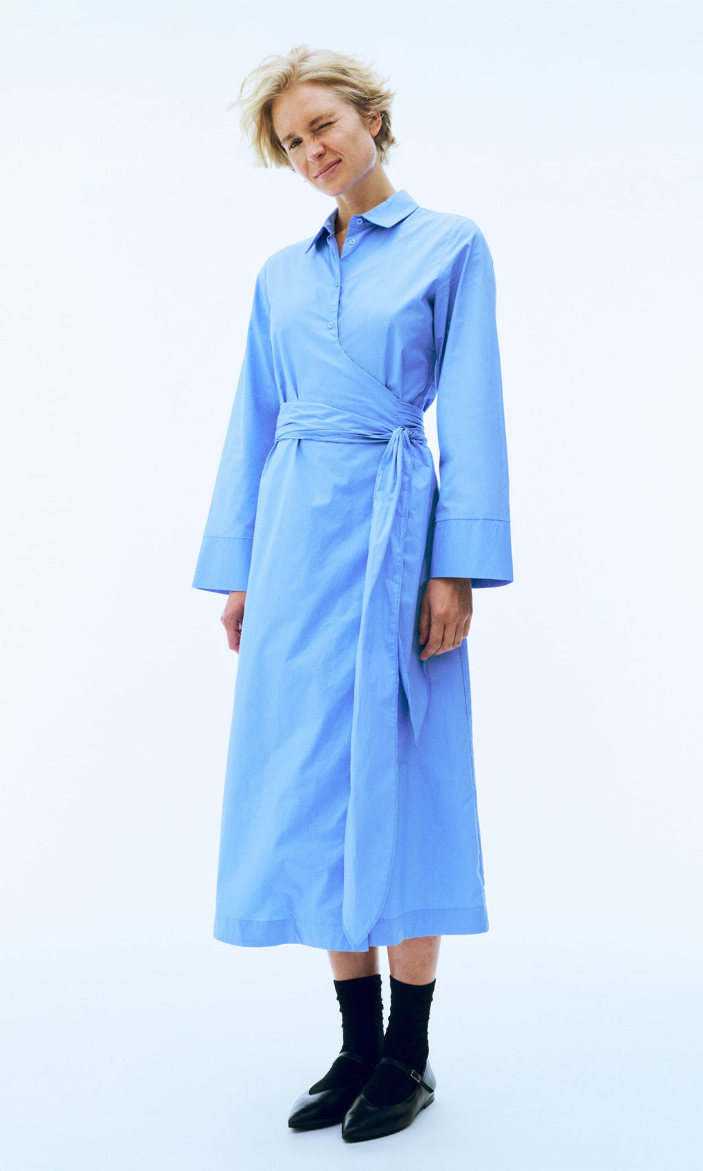 Plümo- Person wearing the Cornflower Dress with a tie belt, black socks, and shoes, standing against a white background.