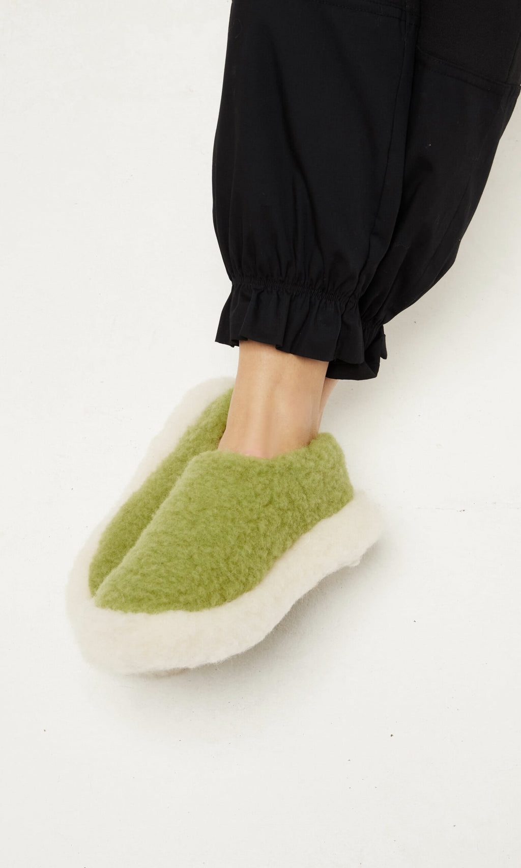 Plümo  A person wearing lime Wool Slippers with white trim and black pants stands on a bright white surface.