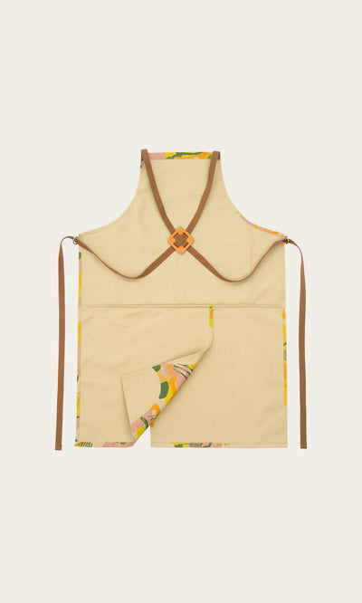 Plümo- The Split Front Apron is beige canvas with brown cross-back straps and a patterned lining at the bottom corner.
