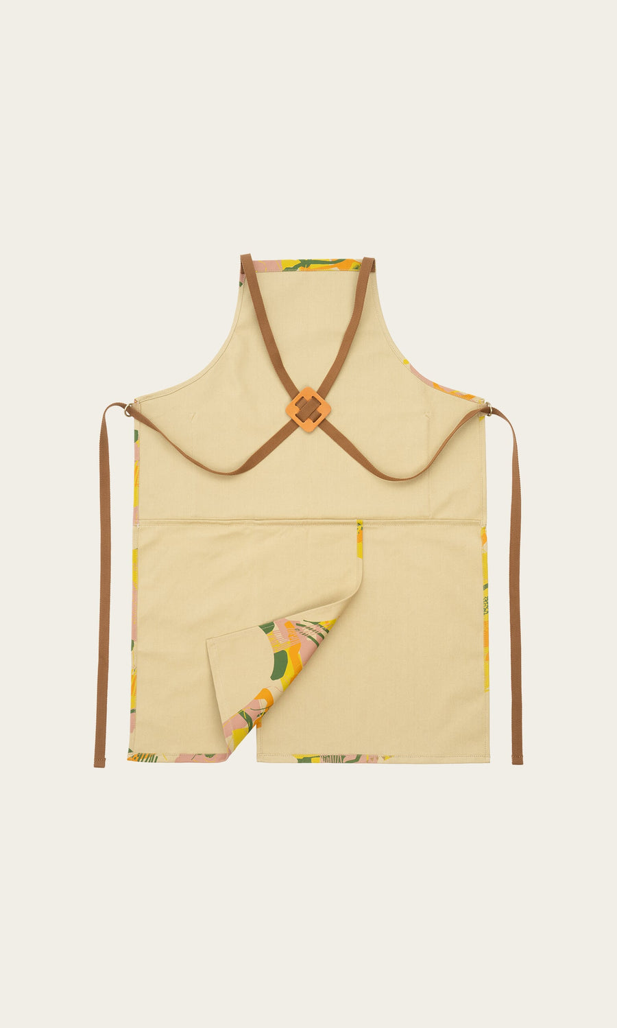 Plümo- The Split Front Apron is beige canvas with brown cross-back straps and a patterned lining at the bottom corner.