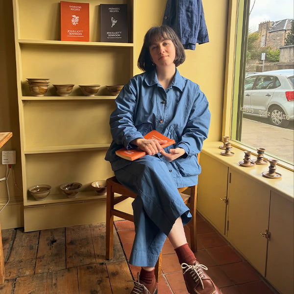 Plümo- A woman in blue clothing sits on a stool by a window, holding a book, with pottery and books on shelves behind her.