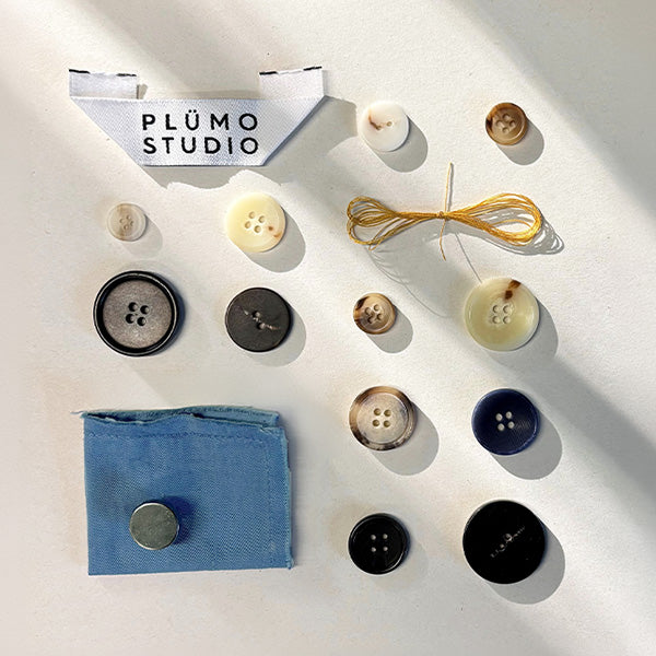 Plümo- Assorted buttons, yellow thread, a blue fabric swatch, and a PLUMO STUDIO label arranged on a white surface.