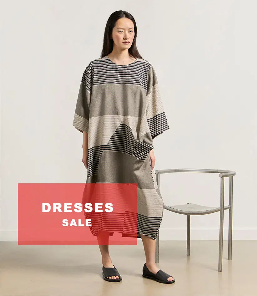 Plümo  Woman in a loose, striped dress stands by a metal chair; red banner reads DRESSES SALE.