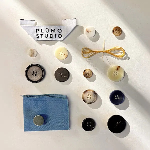 Plümo  Assorted buttons, thread, a fabric swatch, and a PLÜMO STUDIO label arranged neatly on a white surface.