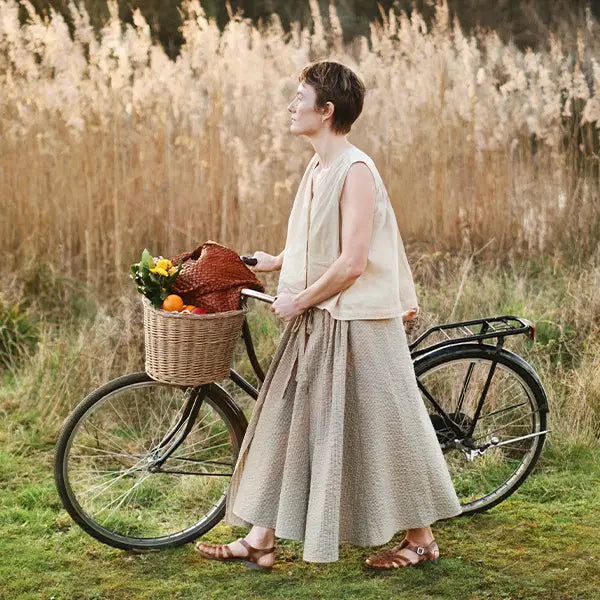 Plümo  A woman in a beige outfit walks a bicycle with a basket of vegetables through a grassy field.