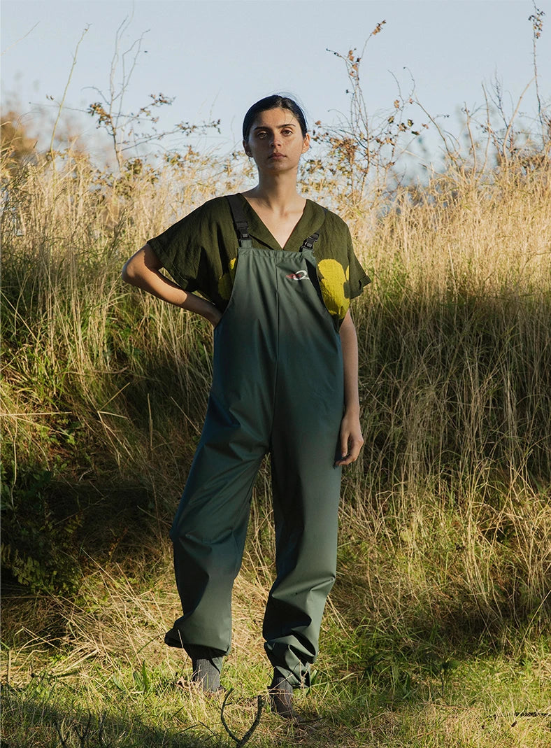 Plümo  A person in green overalls and a patterned shirt stands in tall grass and sunlight outdoors.