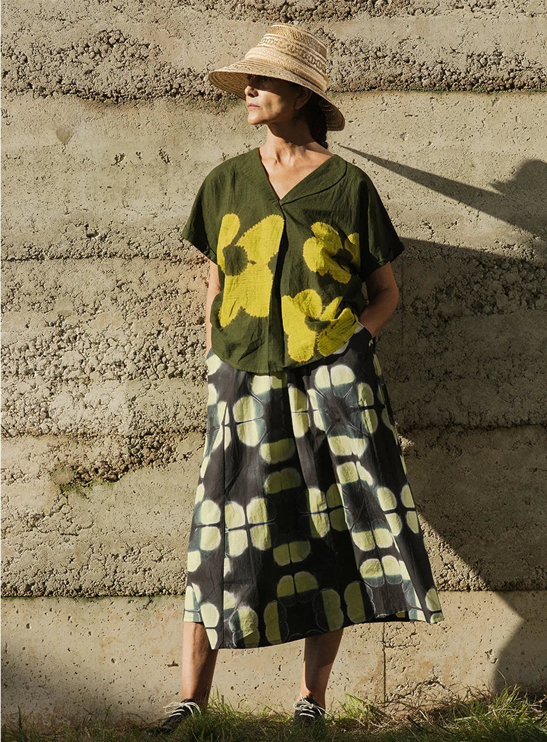 Plümo  A woman in a straw hat wears a green floral top and a black skirt with yellow-green patterns, standing by a textured wall.