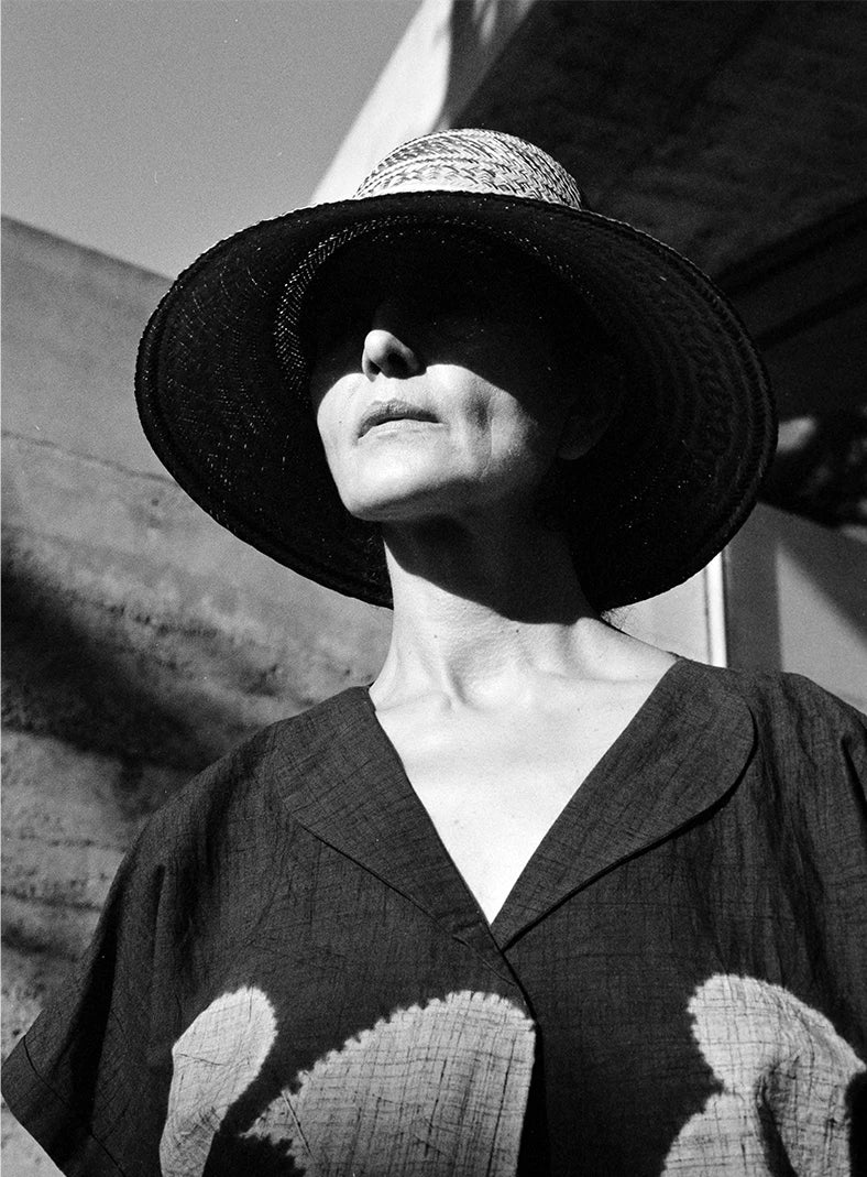 Plümo  Black-and-white photo of a woman in a wide-brimmed hat and patterned shirt, her face partly in shadow.