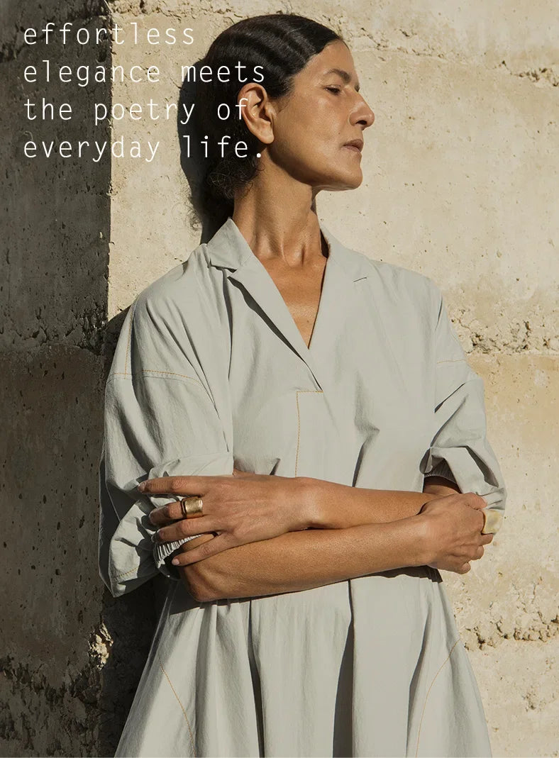 Plümo  A woman in a light dress stands against a sunlit stone wall, arms crossed, with text about elegance and everyday life.
