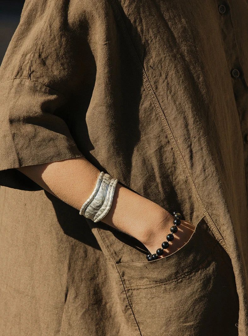 Plümo  A person in a brown linen garment with one hand in a pocket, wearing two bracelets on their wrist.