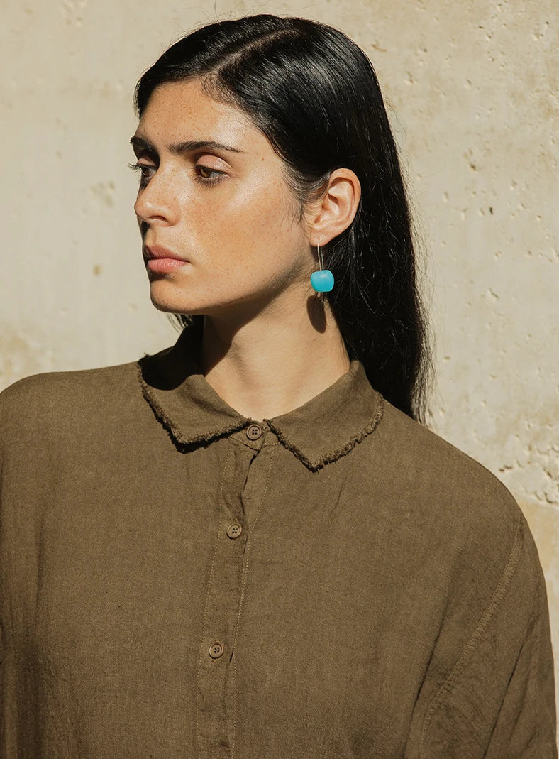 Plümo  Woman with long dark hair, wearing a brown collared shirt and a blue earring, looks to the side in sunlight.