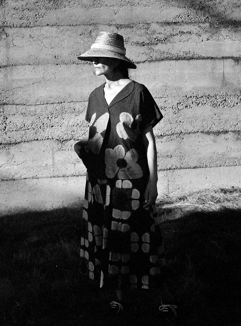Plümo  A woman in a floral dress and wide-brim hat stands in sunlight against a textured concrete wall.