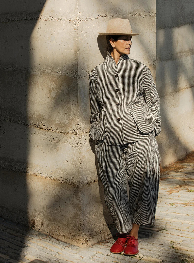 Plümo  Person in a gray textured suit, beige hat, and red shoes leans against a concrete wall in sunlight and shadow.