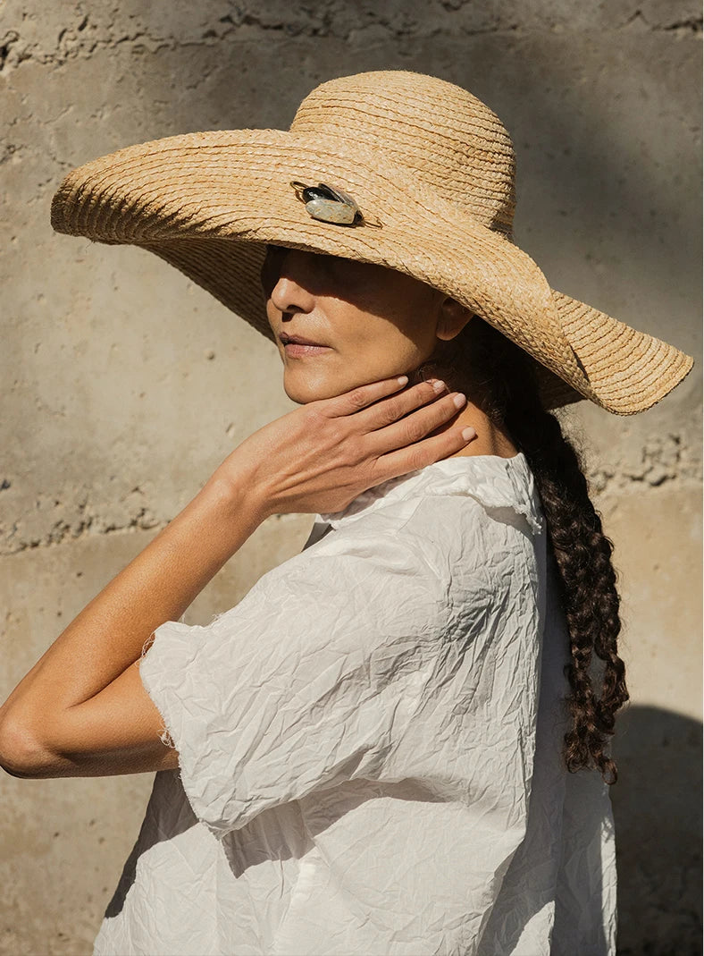 Plümo  Woman in a wide-brimmed straw hat and white shirt, standing against a sunlit textured wall, touching her neck.