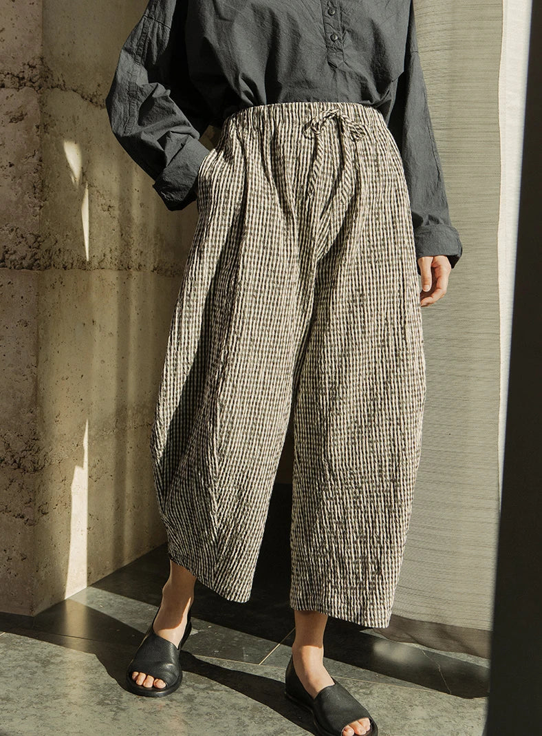 Plümo  Person wearing loose, striped, cropped pants, a dark oversized shirt, and black sandals, standing indoors in sunlight.