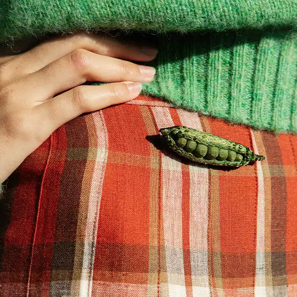Plümo- A hand rests on a red plaid skirt with a green caterpillar cocoon attached, and a green sweater is visible above.