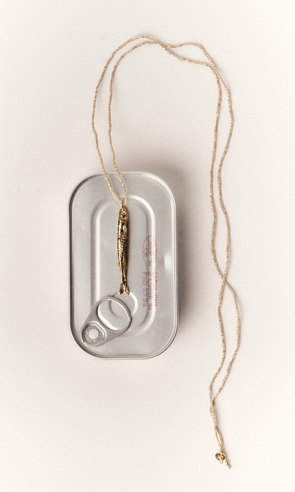 Plümo- The Sardine Necklace, gold-plated, is displayed over a closed silver sardine can on a white background.