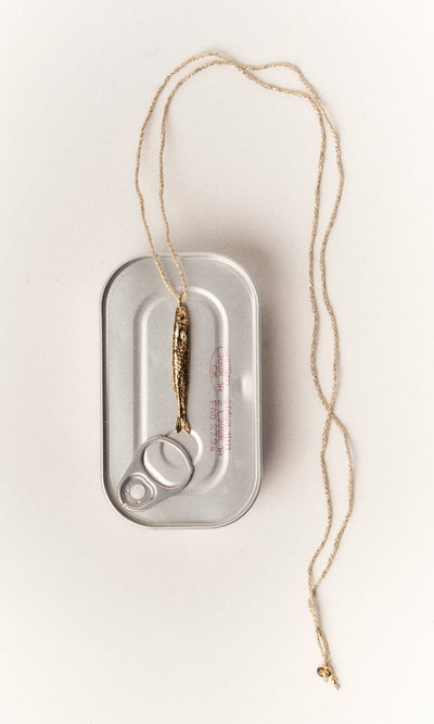 Plümo- The Sardine Necklace, gold-plated, is displayed over a closed silver sardine can on a white background.