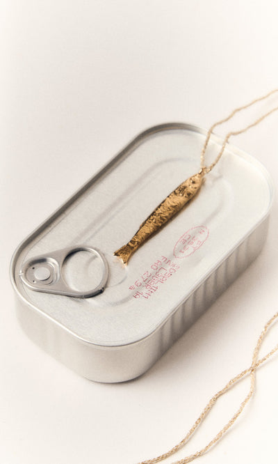 Plümo- The Sardine Necklace features a gold-plated sardine pendant on a chain atop a closed silver tin can.