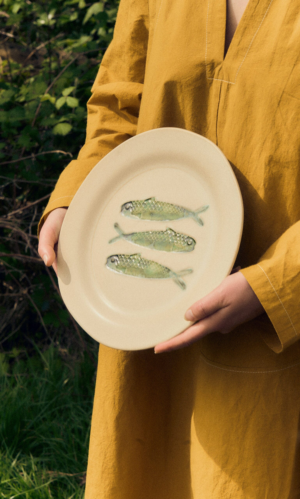 Plümo- A person holds the Sardine Plate outdoors, featuring hand-painted sardines, with greenery in the background.