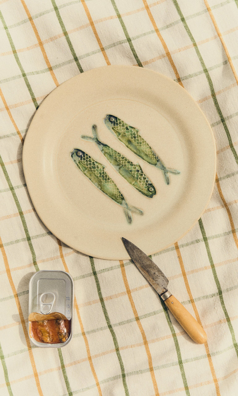 Plümo- The Sardine Plate is a stoneware dinner plate featuring hand-painted sardines, a knife, and an opened tin.