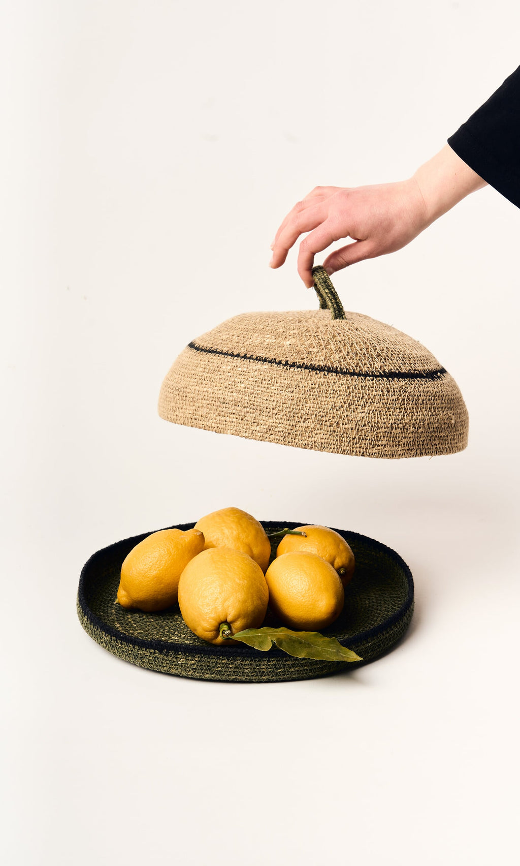 Plümo- A hand lifts the Seagrass Food Cover from a basket of lemons, enhancing rustic decor on a white background.