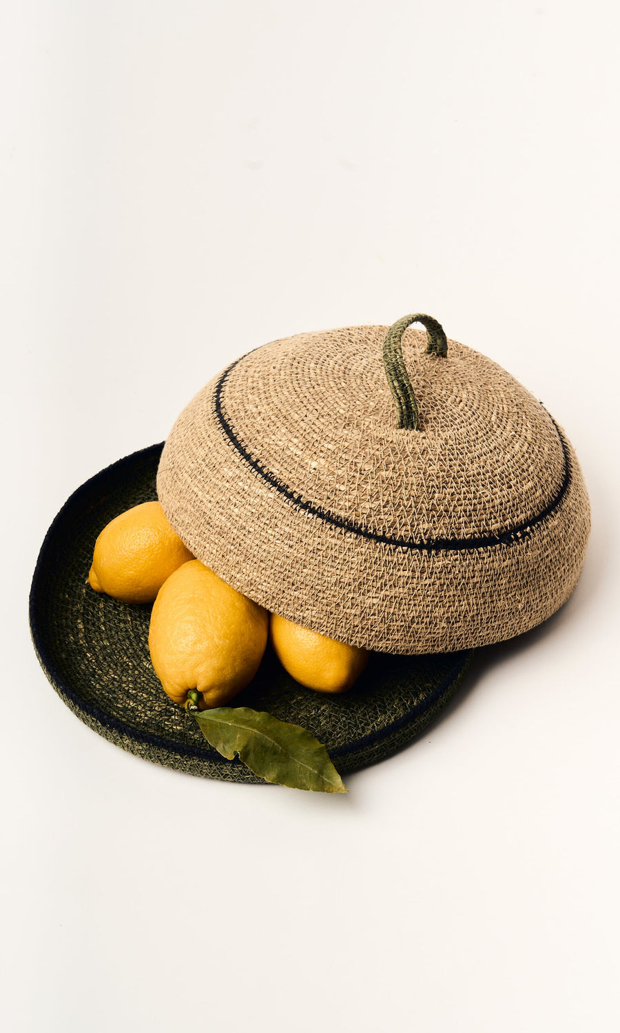 Plümo- Three lemons and a green leaf in a handwoven Seagrass Food Cover, ideal for rustic home decor on a white backdrop.