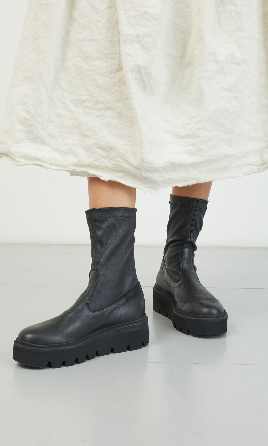 Plümo- Someone wearing Short Prato Boots and a long, off-white skirt stands on a gray floor.