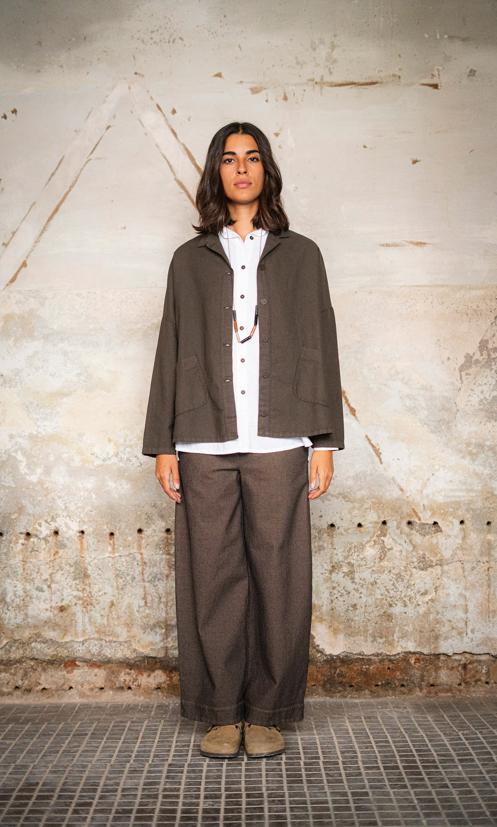Plümo  Woman stands against a textured wall in a boxy Otto Jacket, white shirt, wide pants, and beige shoes.