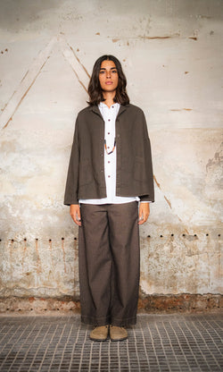 Plümo  Woman stands against a textured wall in a boxy Otto Jacket, white shirt, wide pants, and beige shoes.