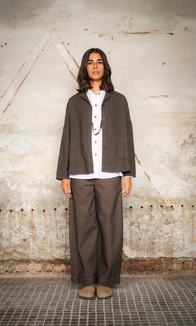 Plümo  Woman stands against a textured wall in a boxy Otto Jacket, white shirt, wide pants, and beige shoes.