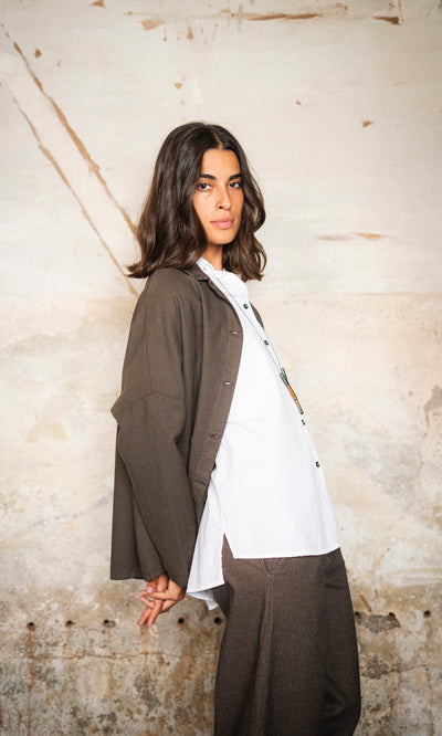 Plümo  Woman with wavy dark hair models the loose-fit brown Otto Jacket against a textured, light-colored wall.