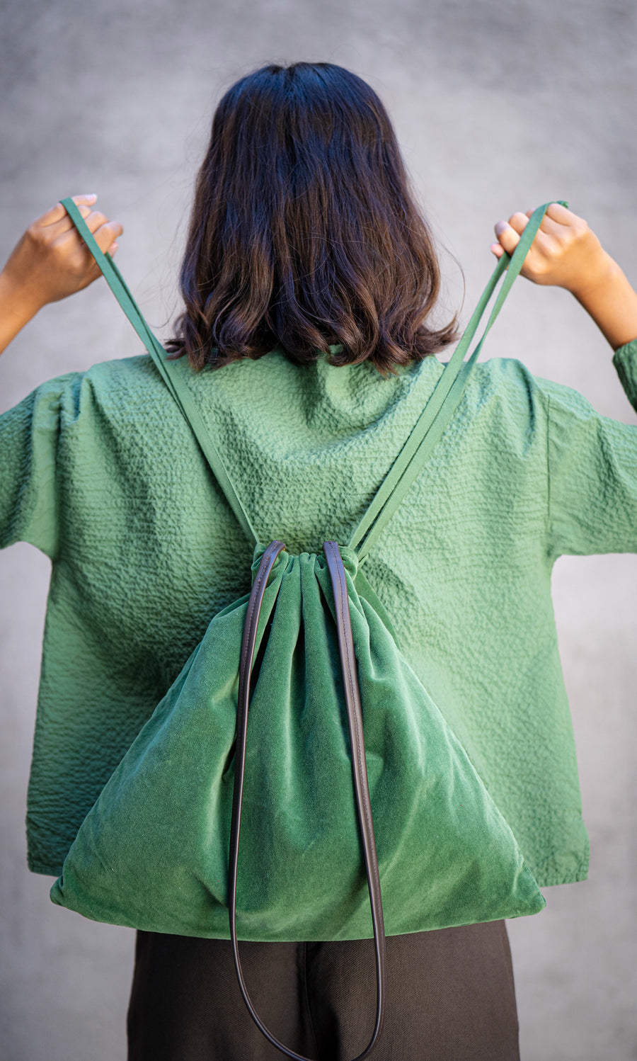 Plümo  Person wears a green textured jacket and holds the Velour Triangle Bag, featuring a drawstring closure, facing away.