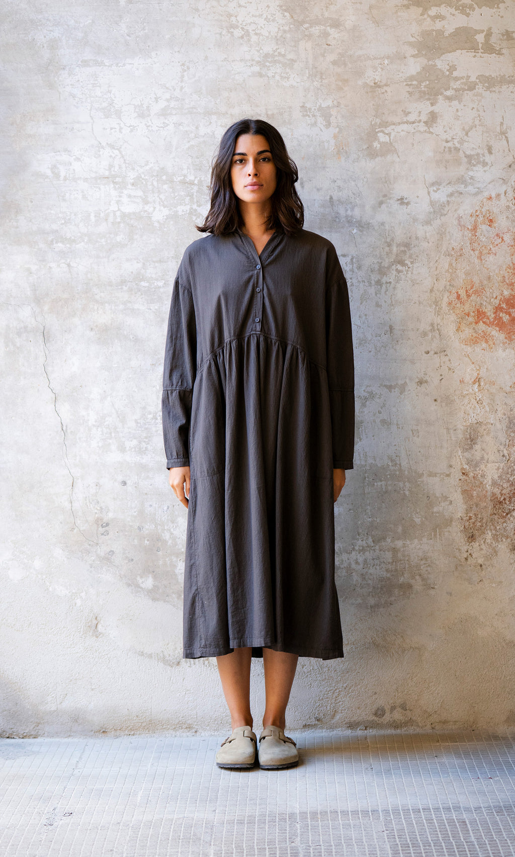 Plümo  Woman wears the Marisol Dress in Truffle, standing against a textured wall and looking straight ahead.