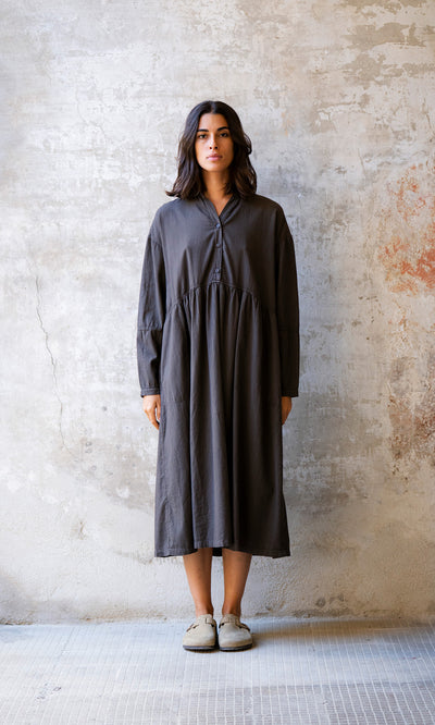 Plümo  Woman wears the Marisol Dress in Truffle, standing against a textured wall and looking straight ahead.