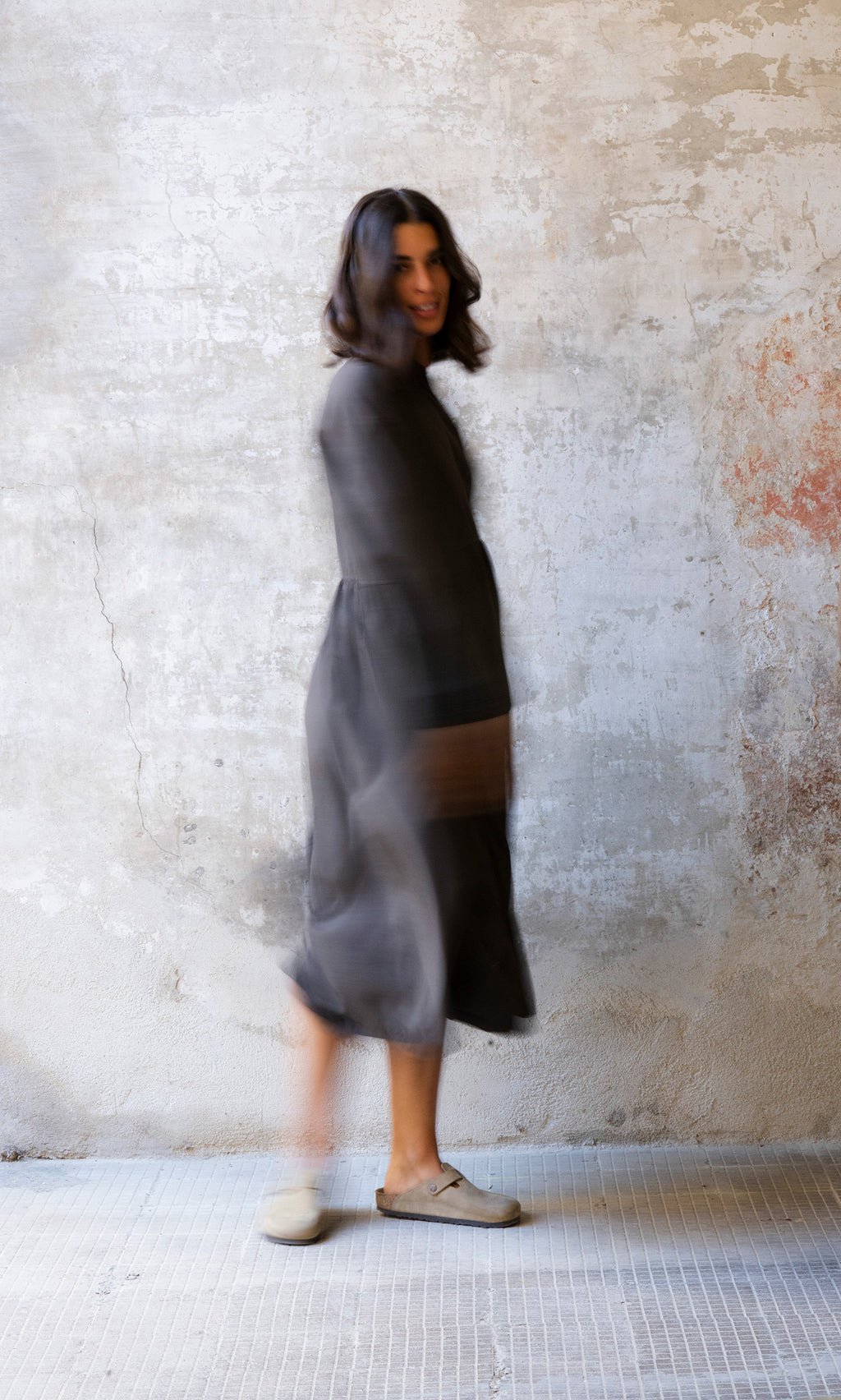 Plümo  Woman in the Marisol Dress - Truffle walks indoors, motion-blurred, against a textured wall in earthy cotton stripes.