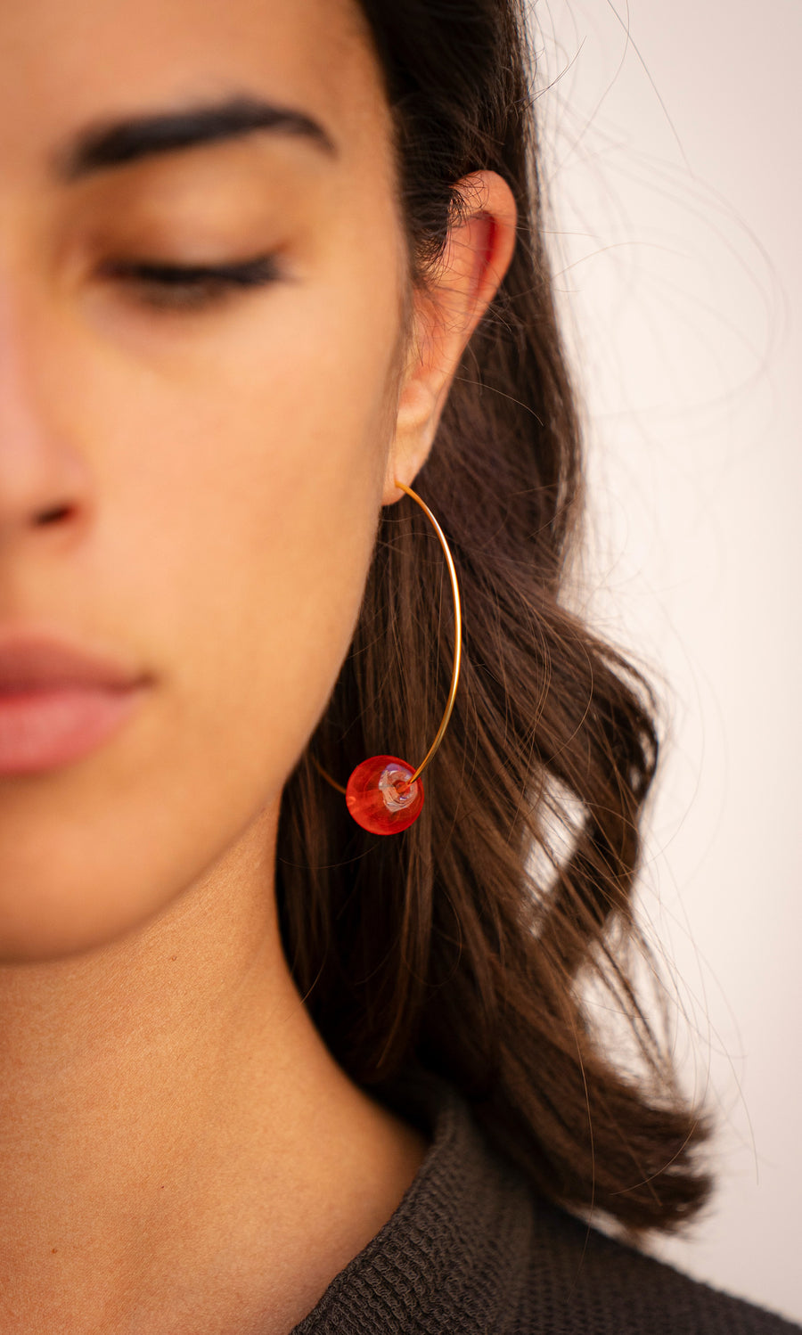 Plümo  Close-up of a woman with closed eyes in Clementine Earrings—gold hoops with a red bead, brown hair visible.