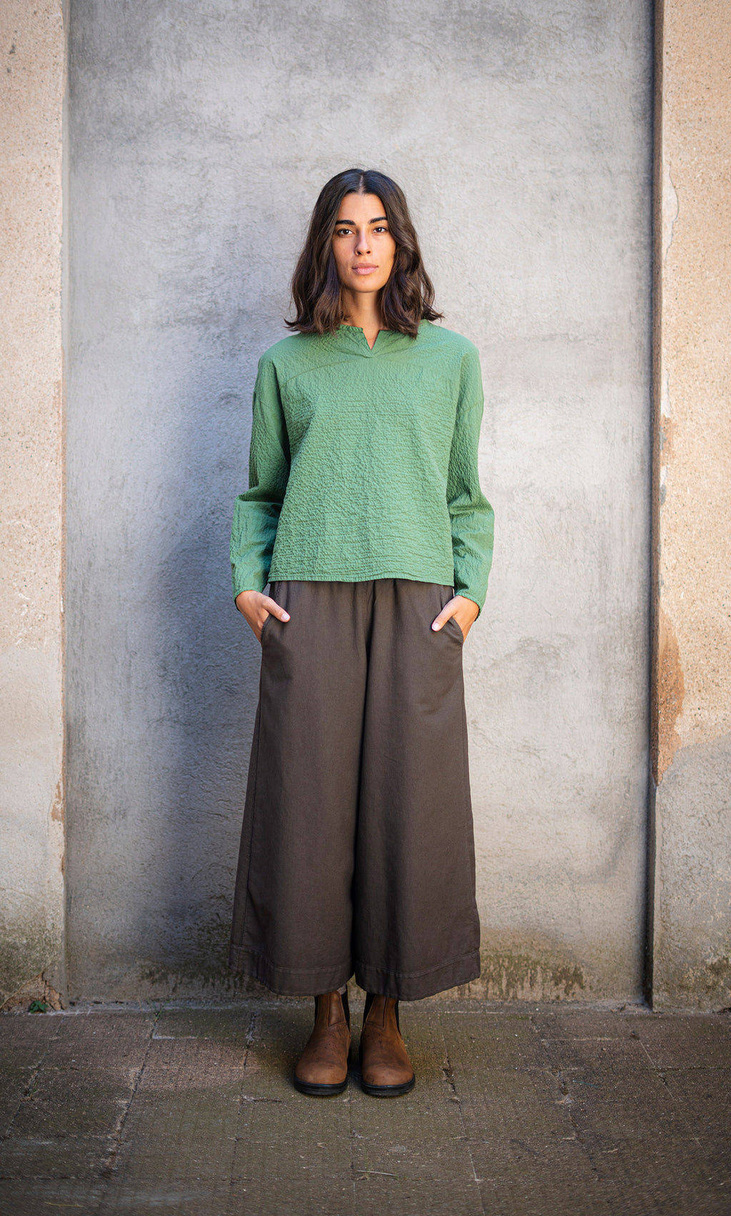 Plümo  Woman in green top and Otto Trousers stands by concrete wall, hands in pockets, looking at camera.