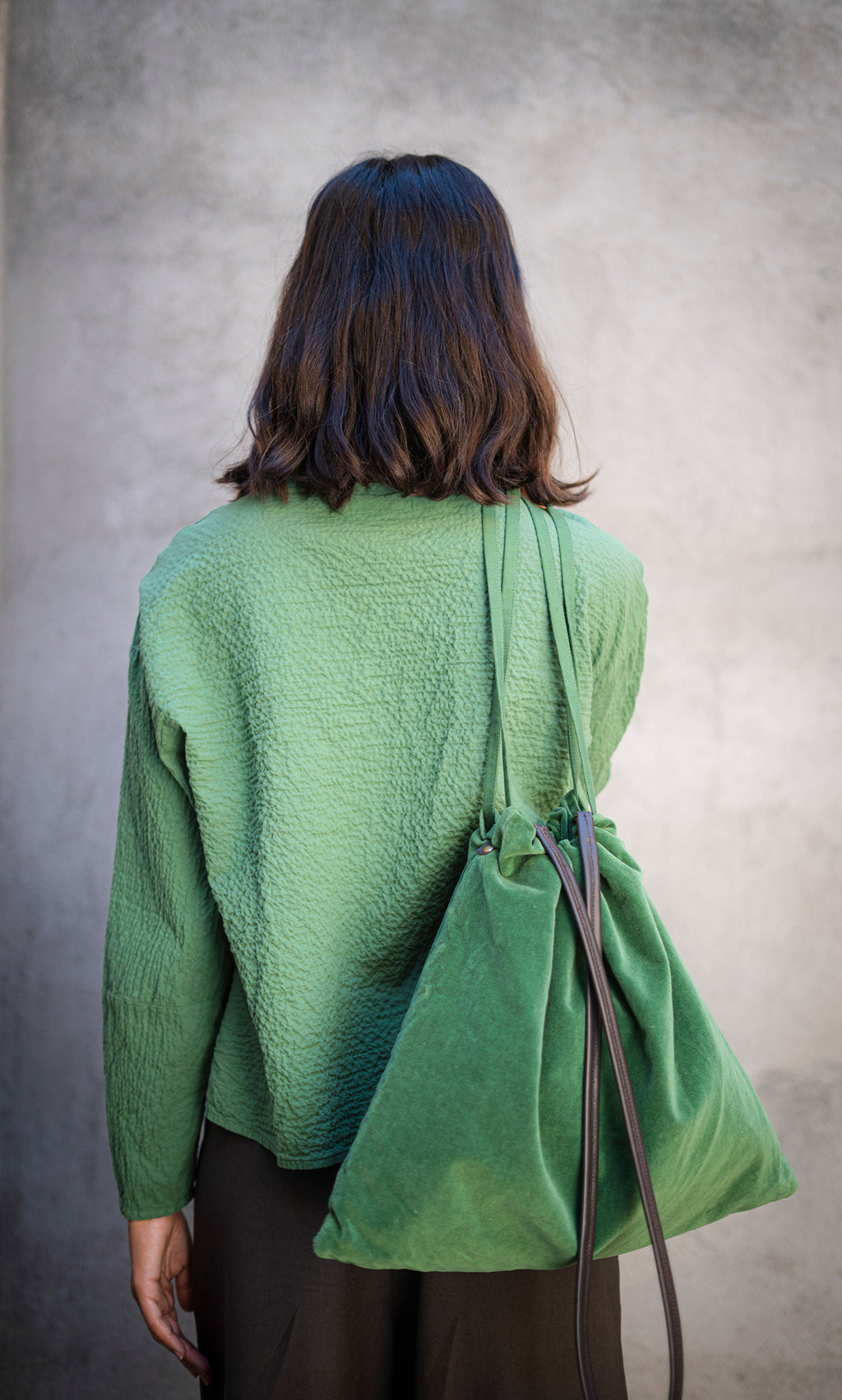 Plümo  Woman with shoulder-length hair in a Verde Blouse and matching bag, seen from behind against a gray wall.