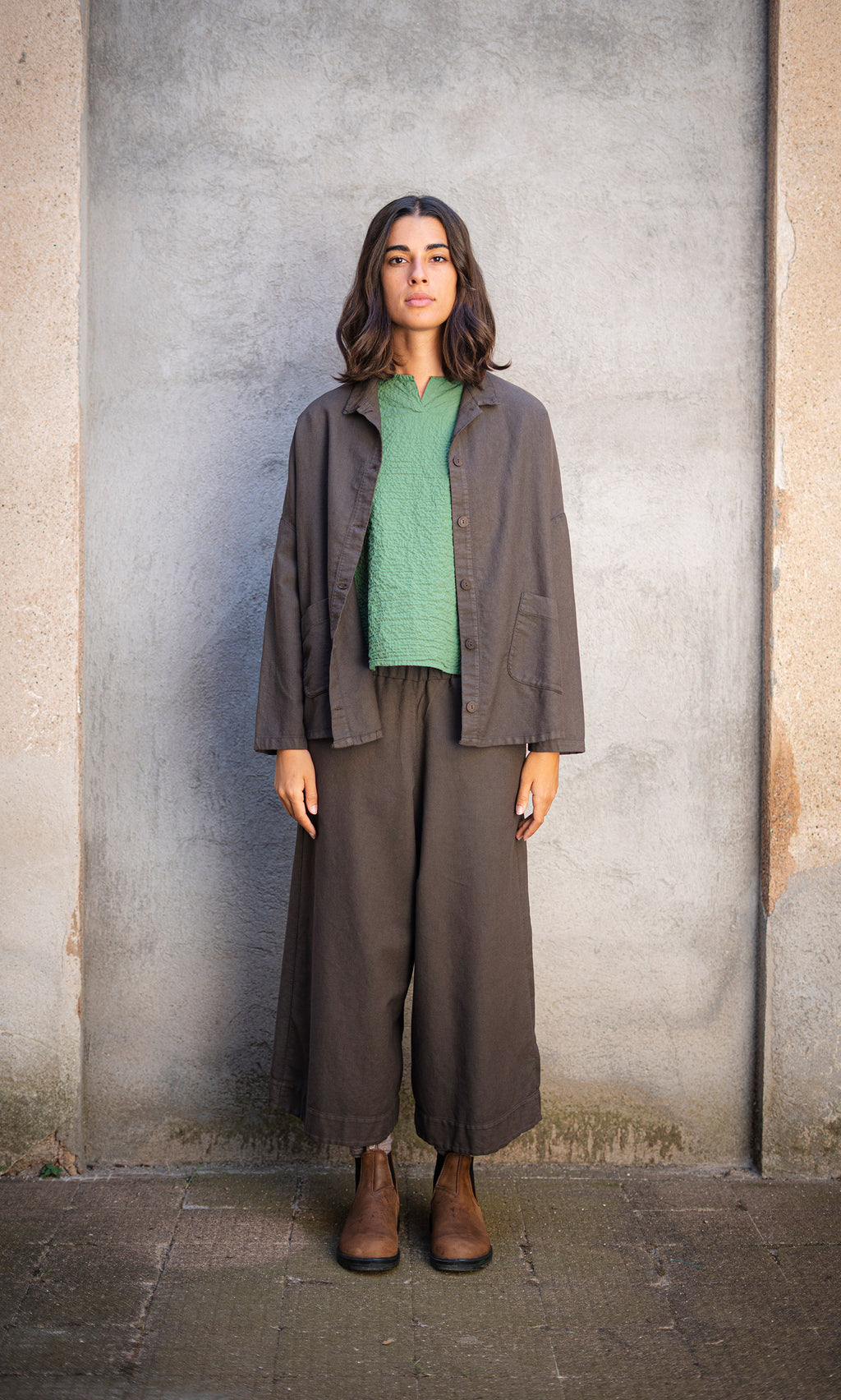 Plümo  Person with shoulder-length hair stands against a gray wall, wearing Otto Trousers and workwear boots.